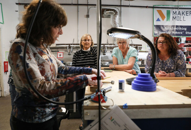 Deb Ryan (left) leads a class in the metalsmithing shop at Makers Mill, demonstrating how to use a flex shaft to drill holes in metal jewelry. In this class the students learn how to cut and texture silver, brass and copper metal to make pendants and earrings. (Photo by Cheryl Senter.)