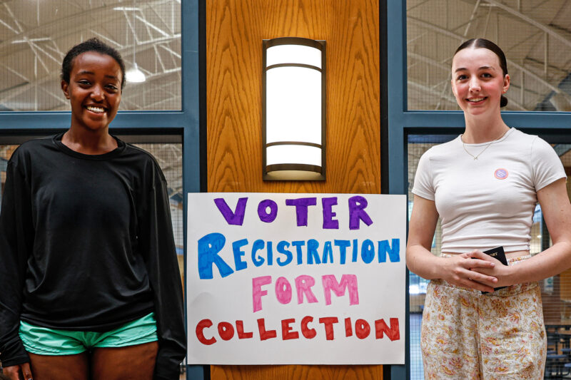 Two high school students stand with a "Voter registration form collection sign" in their school