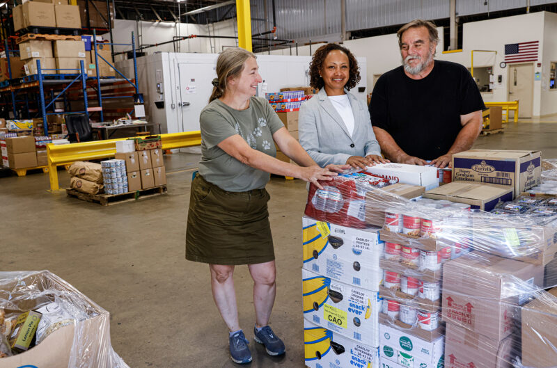 Three people stand with a palette of food in a food bank warehouse